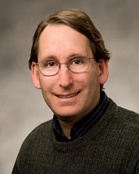 Head shot of Dr. Joel Sipress, a white male. Joel is wearing glasses and a sweater and smiling. The background is blurred.